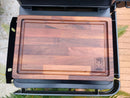 The BBQ Boards® Traeger Timberline XL Side Cutting Board sits on a black grill prep area, featuring the BBQ Boards® logo with surrounding greenery and paving in the background.