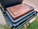 The BBQ Boards® Traeger Timberline XL Side Cutting Board by BBQ Boards® is placed on a grill prep area outdoors atop a closed Traeger Timberline XL side board, with brick paving and green plant leaves in the background.