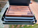 The BBQ Boards® Traeger Timberline XL Side Cutting Board by BBQ Boards® sits on a metal shelf attached to a black outdoor grill, creating an ideal prep space with brick pavement and green plants in the background.