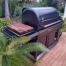 A BBQ Boards® Traeger Timberline XL Side Cutting Board by BBQ Boards® rests on the prep space of a large black outdoor grill with a closed lid, situated on a brick patio surrounded by lush greenery, flowers, and trees.