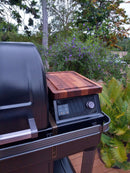 A BBQ Boards® Traeger Timberline XL Pellet Bin Cutting Board sits on the side shelf of a Traeger Timberline XL grill with a closed lid, while green plants and purple flowers brighten the background.