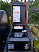 A close-up of the BBQ Boards® Traeger Timberline XL Pellet Bin Cutting Board by BBQ Boards®, shown on a grill with the hopper lid open, empty pellet container, instruction labels, and green plants in the background.