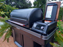 A BBQ Boards® Traeger Timberline XL Pellet Bin Cutting Board by BBQ Boards® is placed on a modern black outdoor grill with a closed lid and an open side compartment, set on a patio surrounded by green plants and trees.