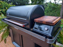 A BBQ Boards® Traeger Timberline XL Pellet Bin Cutting Board by BBQ Boards®, shown atop a modern outdoor Traeger Timberline XL grill with a closed lid, digital panel, and side shelf, on a patio surrounded by lush plants.