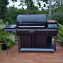 A Traeger Timberline XL grill with a closed lid is on a brick patio among greenery, featuring side shelves and a BBQ Boards® Traeger Timberline XL Pellet Bin Cutting Board by BBQ Boards® on the right.
