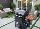A BBQ Boards® Weber Searwood 600 Pellet Bin Cutting Board rests on a modern black grill beside a wooden side table on the patio, surrounded by wicker furniture, green artificial grass strips, potted plants, and sliding glass doors.