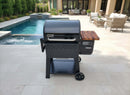 A black Weber Searwood 600 grill with a wooden side shelf sits on a light stone patio by the pool, surrounded by patio furniture and greenery, featuring the BBQ Boards® Weber Searwood 600 Pellet Bin Cutting Board in the background.