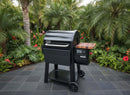 A Weber Searwood 600 grill with a BBQ Boards® Pellet Bin Cutting Board stands on a tiled patio, surrounded by lush green palm trees and vibrant tropical plants.