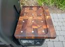 The BBQ Boards® Weber Searwood 600 Pellet Bin Cutting Board, a thick rectangular magnetic board with a checkered pattern and rivets, sits on the Weber Searwood 600’s side shelf above the digital temperature display, with paving stones and green plants in view.