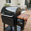 A black Weber Searwood 600 grill, featuring a BBQ Boards® Weber Searwood 600 Pellet Bin Cutting Board and digital control panel, stands on a wooden patio deck surrounded by outdoor furniture and plants.