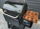 A black Weber Searwood 600 pellet grill with a shiny lid, stainless steel handle, and the BBQ Boards® Weber Searwood 600 Pellet Bin Cutting Board by BBQ Boards® attached to the side. A digital display shows temperature readings.