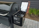 A BBQ Boards® Weber Searwood 600 Pellet Bin Cutting Board is shown on a patio with gray pavers, beside green plants, placed near a grill with a digital temperature panel and an open side compartment displaying instructions.