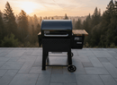 A black Weber Searwood 600 grill with a wood side shelf sits on a stone patio at sunset, overlooking forested views. A BBQ Boards® Weber Searwood 600 Pellet Bin Cutting Board from BBQ Boards® adds extra prep space.