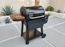 A black Weber pellet grill with a wooden side shelf and digital panel stands on gray patio stones, featuring the BBQ Boards® Weber Searwood 600 Side Cutting Board for easy prep, with desert plants in the background.