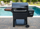 A BBQ Boards® Weber Searwood 600 Side Cutting Board rests on the side prep area of a Weber grill, set on a stone patio by the pool with green plants and lounge chairs in the background.