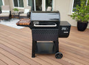 A black Weber pellet grill with a BBQ Boards® Weber Searwood 600 Side Cutting Board by BBQ Boards® sits on a wooden deck, featuring a digital control panel and wheels, with patio furniture and potted plants in the background near a house.
