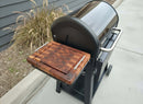 A black outdoor grill with a BBQ Boards® Weber Searwood 600 Side Cutting Board attached to its side shelf sits on a concrete patio near ornamental grasses and a dark-paneled wall.