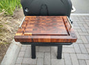 The BBQ Boards® Weber Searwood 600 Side Cutting Board, with metal handles and a carved groove, sits on a grill side shelf. Paving stones and green plants in the background make it ideal for outdoor pellet grill prep.