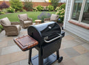 A black grill with a BBQ Boards® Weber Searwood 600 Side Cutting Board stands on a stone patio. Wicker chairs and a small table complete the cozy area overlooking a lush backyard garden with grass and flowers.