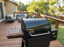 A black Weber smoker grill sits on a wooden deck with outdoor sofas, a table, and green trees in the background, featuring the BBQ Boards® Weber Searwood 600 Side Cutting Board ready for pellet grill prep on a sunny day.