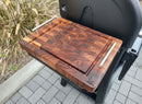 The BBQ Boards® Weber Searwood 600 Side Cutting Board, featuring an end-grain pattern and carved logo, sits on a grill’s side shelf. Metal handles add style while pavers and ornamental grass are visible nearby.