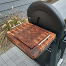 The BBQ Boards® Weber Searwood 600 Side Cutting Board, made from dark end-grain wood with stainless steel handles, rests on a black grill’s side shelf. A bush behind it creates an ideal pellet grill prep space.