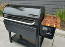 A black Weber Searwood XL 600 pellet grill with a digital control panel and wooden side shelf stands outdoors on concrete, accompanied by the BBQ Boards® Weber Searwood XL 600 Pellet Bin Cutting Board for convenient storage and prep.