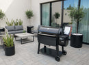 A modern patio features a Weber Searwood XL 600 pellet grill, gray wicker furniture, a coffee table, potted plants, and a BBQ Boards® Weber Searwood XL 600 Pellet Bin Cutting Board on light gray pavers beside large windowed building.
