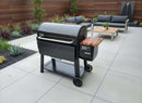 A black Weber Searwood XL 600 pellet grill with a wooden side shelf sits on a modern patio with outdoor furniture and plants. A BBQ Boards® Weber Searwood XL 600 Pellet Bin Cutting Board is ready for prep nearby.