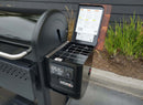 A Weber Searwood XL 600 outdoor grill with a digital temperature panel and a recipe guide inside the lid sits next to a BBQ Boards® Weber Searwood XL 600 Pellet Bin Cutting Board, all arranged on paving by tall grass and a dark building.