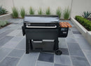 A black Weber Searwood XL 600 grill with a BBQ Boards® Weber Searwood XL 600 Pellet Bin Cutting Board rests on a stone patio, surrounded by neat plants and tall grasses beside a light gray wall.