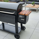 A BBQ Boards® Weber Searwood XL 600 Pellet Bin Cutting Board sits on a table next to a black pellet grill on a modern patio with gray tiles, surrounded by outdoor furniture and a small potted plant.