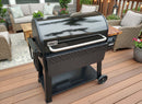 A BBQ Boards® Weber Searwood XL 600 Pellet Bin Cutting Board is displayed on a wooden deck with wicker patio furniture, potted plants, and outdoor lighting for a cozy backyard atmosphere.