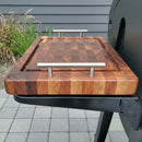 A BBQ Boards® Weber Searwood XL 600 Side Cutting Board with metal handles, checkered pattern, and a juice groove sits on a grill’s side shelf, making it an ideal BBQ accessory. Outdoor tiles and plants can be seen in the background.