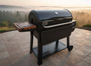 A BBQ Boards® Weber Searwood XL 600 Side Cutting Board by BBQ Boards® is attached to a black Weber grill on a stone patio, overlooking a misty forest at sunrise.
