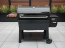 A black Weber pellet grill with a BBQ Boards® Weber Searwood XL 600 Side Cutting Board and digital control panel stands on a tiled patio, surrounded by tall green plants and wooden fencing.