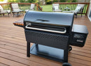 A black Weber pellet grill is on a wooden deck next to the BBQ Boards® Weber Searwood XL 600 Side Cutting Board. Outdoor patio furniture and green trees are in the background, and the grill’s digital display shows 275°F.