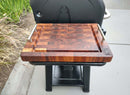 The BBQ Boards® Weber Searwood XL 600 Side Cutting Board with metal handles sits on a black outdoor grill’s side shelf, offering a stylish BBQ side board on a concrete patio, with green plants as a lush backdrop.