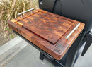The BBQ Boards® Weber Searwood XL 600 Side Cutting Board, featuring metal handles and a rich patterned grain with an engraved logo, rests on a black grill side shelf outdoors near ornamental grasses and a curb.