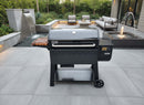 A BBQ Boards® Weber Searwood XL 600 Side Cutting Board is featured with a black Weber pellet grill on a modern gray patio, surrounded by outdoor couches, a water feature, and lush greenery.