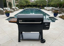 A black Weber grill and a BBQ Boards® Weber Searwood XL 600 Side Cutting Board are set on a tiled patio by a rectangular pool, surrounded by outdoor seating, potted plants, and palm trees in a landscaped backyard.