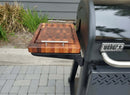 A BBQ Boards® Weber Searwood XL 600 Side Cutting Board with metal handles sits on the shelf of a black grill, serving as an ideal BBQ side board on the patio near plants.
