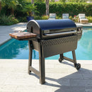 A BBQ Boards® Weber Smoque & XL Side Cutting Board by BBQ Boards® is featured on a black outdoor grill, set on a stone patio next to a pool and surrounded by greenery and lounge chairs.