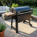 A BBQ Boards® Weber Smoque grill with an XL Side Cutting Board stands on a stone patio, surrounded by potted plants, outdoor furniture, and a lush green lawn in the backyard.