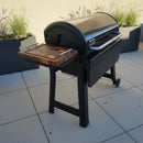 A black pellet grill prep station with a BBQ Boards® Weber Smoque & XL Side Cutting Board stands on a tiled patio, framed by decorative planters of tall grasses in the background.