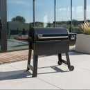 A BBQ Boards® Weber Smoque & XL Side Cutting Board from BBQ Boards® is paired with a black outdoor pellet grill on a concrete patio before glass doors, with plants and outdoor furniture in the background.