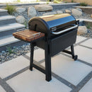 A black outdoor grill with a curved lid and side handle features the BBQ Boards® Weber Smoque & XL Side Cutting Board by BBQ Boards®, standing on a modern patio with stone pavers and decorative rocks in the background.