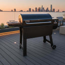 A black Weber grill and a BBQ Boards® Weber Smoque & XL Side Cutting Board are set on a rooftop deck with modern seating, potted plants, and city skyscrapers in the sunset background.