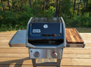 A Weber gas grill with a closed lid and two control knobs stands on a wooden deck with a side shelf, featuring a BBQ Boards® Weber Spirit II Side Cutting Board. Trees and greenery fill the background.