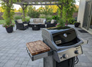A modern patio features a gas grill with a BBQ Boards® Weber Spirit Old Gen Side Cutting Board, wicker outdoor furniture and cushions around a fire pit, all surrounded by greenery and potted plants.
